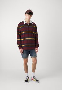 Bordo svītrains garām piedurknēm polo krekls ar baltu apkakli, apvienots ar džinsu šortiem. Melni un dzelteni rakstaini sporta apavi ar baltiem zeķēm. Brūna cepure.