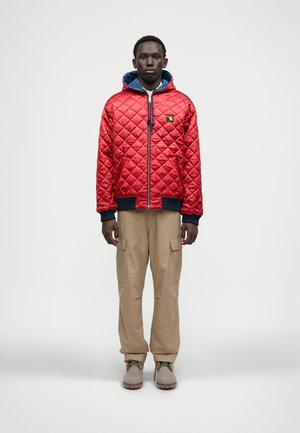 Chaqueta acolchada roja con capucha azul, cierre de cremallera, puños acanalados, combinada con pantalones cargo beiges y botas grises.