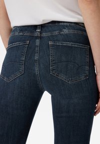 Jeans de denim azul oscuro con un diseño ajustado, que cuentan con dos bolsillos traseros y costuras en contraste a lo largo de las costuras. Textura suave en general.