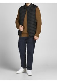 Czarna pikowana kamizelka na brązowym, teksturowanym swetrze, zestawiona z niebieskimi dżinsami i białymi trampkami. Posiada wysoki kołnierz i zamek błyskawiczny.