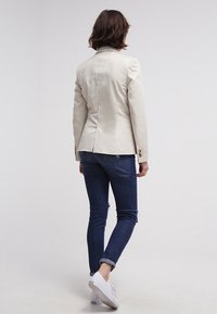 Femme aux cheveux bruns portant un blazer crème, un jean bleu foncé et des baskets blanches, debout contre un fond blanc uni, dos tourné vers le spectateur.