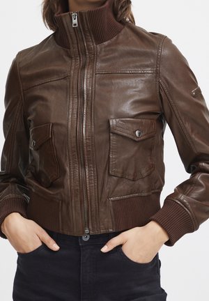 Gros plan sur une personne portant une veste en cuir marron foncé zippée avec poignets côtelés, ceinture, col montant et deux poches avant à rabat avec boutons.