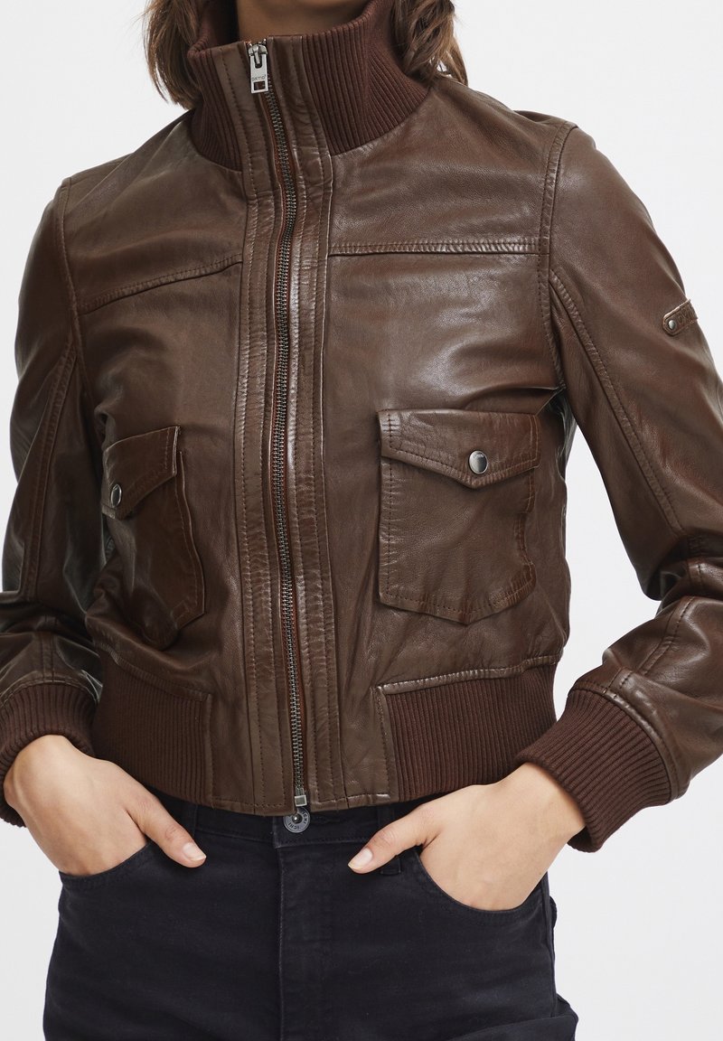 Gros plan sur une personne portant une veste en cuir marron foncé zippée avec poignets côtelés, ceinture, col montant et deux poches avant à rabat avec boutons.