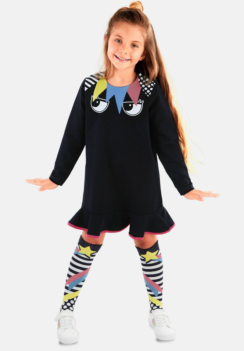 Vestido negro con un dobladillo con volantes, que presenta un diseño gráfico colorido en el cuello. Combinado con calcetas de rodilla de estampado y zapatos blancos.