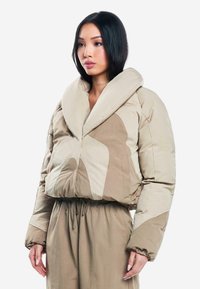 Abrigo tipo plumas beige con cuello oversized, patrones texturizados y cinturilla elástica. Lleva encima un conjunto a juego de color beige.