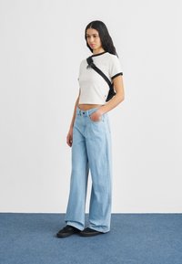 Γυναίκα με μακριά σκούρα μαλλιά που φοράει λευκό κοντό t-shirt, ανοιχτό μπλε τζιν με φαρδιά μπατζάκια, μαύρα sneakers και μαύρη τσάντα χιαστί.