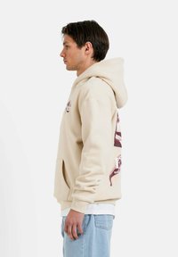 Beige hoodie med känguruficka, med röda grafiska mönster på ryggen och vänster bröst, tillverkad av mjukt, texturerat tyg.