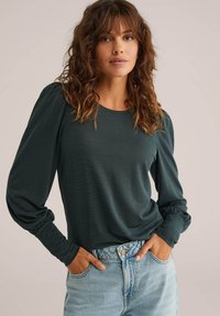 Sötétzöld, hosszú ujjú felső puffos vállakkal és ráncolt mandzsettával, magas derekú, világoskék farmernadrággal párosítva. Simább szövettextúra.