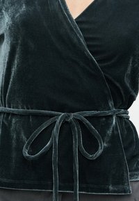 Tumma vihreä samettinen kietaisupaita, jossa on V-pääntie, solmittava vyötärö ja sileä tekstuuri. Ominaisuuksiin kuuluu virtaava muotoilu ja hienovarainen kiilto.