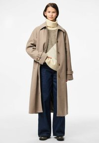 Lungo cappotto beige di lana con colletto, bottoni frontali e tasche laterali, indossato sopra un dolcevita crema e pantaloni a gamba larga in denim scuro.