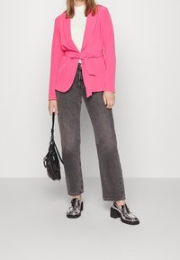 Blazer fuchsia avec taille ceinturée, associé à un jean large gris et des mocassins argentés brillants. Le mannequin tient un petit sac à main noir.