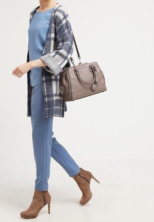 Vrouw met blauwe outfit, geruit vest, bruine suède enkellaarsjes, die een taupekleurige leren handtas over haar schouder draagt terwijl ze loopt.