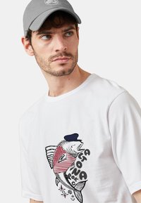 Camiseta de algodón blanca con un gráfico de pez colorido con un diseño de rayas, que lleva puesta una gorra azul marino. La gorra es gris con un logotipo redondo.