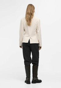Blazer ajustado de color beige claro con una abertura en la parte posterior, combinado con pantalones oscuros de rayas y botas de cuero marrón hasta la rodilla.