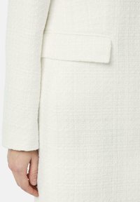 Weißer strukturierter Blazer aus gewebtem Material, mit einer einzelnen Tasche und einem geraden Ärmeldesign, in einer Nahaufnahme gezeigt.