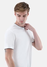 Weißes kurzärmeliges T-Shirt mit schwarzem Kragen. Es hat eine glatte Textur und einen taillierten Schnitt. Wird von einer Person getragen, die den Kragen richtet.