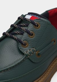 Πράσινο δερμάτινο παπούτσι τύπου boat με σκούρες μπλε κορδόνια με σχέδιο, ορειχάλκινα κορδόνια με τη χαραγμένη λέξη "Tommy Jeans" και ένα λογότυπο κόκκινου, λευκού και μπλε χρώματος στη γλώσσα του παπουτσιού.