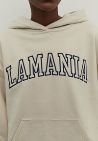 Tejbézs színű kapucnis pulóver, nagy sötétkék hímzett "LAMANIA" felirattal. Kanguru zsebbel és húzózsinórral ellátott kapucnival rendelkezik. Simább textúrájú.
