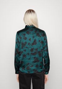 Grüne Satinbluse mit schwarzem Blumenmuster, langen Ärmeln und einem Kragen. Glatte Textur mit subtiler Helligkeit. Taillierte Passform an der Taille.