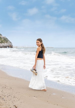 Meisje in een marineblauwe tanktop en een witte rok met stippen, die sandalen vasthoudt, terwijl ze langs een zandstrand aan de zee loopt.