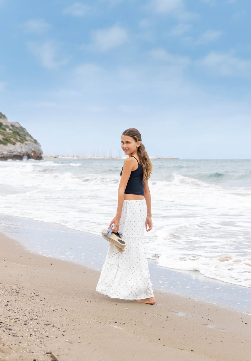 Dziewczyna w granatowej bluzce bez rękawów i białej spódnicy w kropki, trzymająca sandały, spacerująca wzdłuż piaszczystej plaży nad morzem.