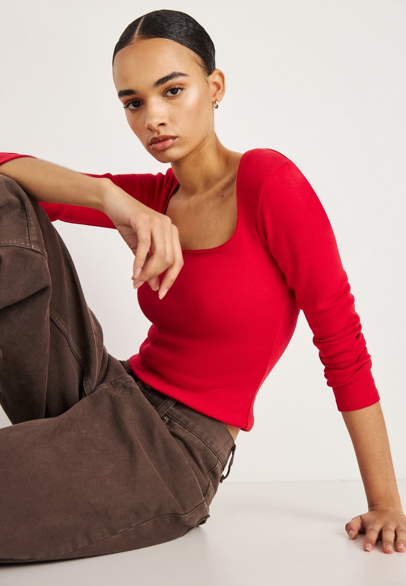 Crvena rebrasta majica s dugim rukavima i kvadratnim izrezom na vratu, uparena s hlačama smeđe boje visokog struka. Glatka tekstura i uski kroj ističu siluetu.