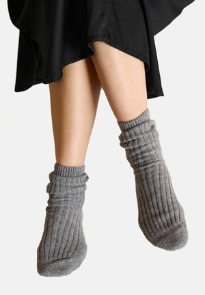 Sive rebraste nogavice z ohlapnim dizajnom, s širokim robom in mehko teksturo, delno vidne nad črnim tekstilom.
