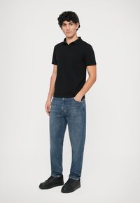 Νεαρός άνδρας με σγουρά μαλλιά, φορώντας μαύρο πόλο πουκάμισο, μπλε τζιν και μαύρα αθλητικά παπούτσια, να στέκεται με το ένα χέρι στην τσέπη, μπροστά σε λευκό φόντο.