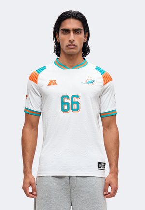 Mężczyzna o ciemnych włosach nosi biały jersey futbolowy Miami Dolphins z numerem 66 i szare spodnie dresowe, stoi na jednolitym tle.