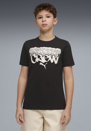 Niño con camiseta negra con texto estilo graffiti "Street Crew" y logotipo de Puma, combinado con pantalones cortos beige claro, de pie contra un fondo gris.