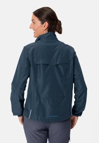 Mørkeblå vanntett jakke med ståkrage, baklommepose og sideslits. Har en glatt tekstur og logo på nederste del av ryggen.