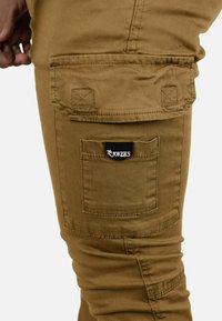 Pantalones cargo color caqui de corte ajustado, con un bolsillo lateral, costuras reforzadas y una parche negro con el logo cerca del bolsillo.