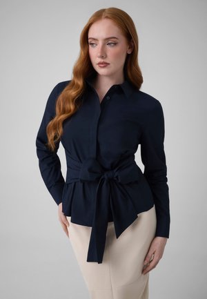 Femme aux longs cheveux roux portant un chemisier bleu marine noué à la taille avec un grand nœud et une jupe beige, posant devant un fond gris.