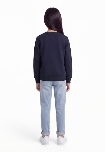 Barn med langt mørkt hår iført marineblå genser, lyseblå oppbrettede jeans og hvite joggesko, står vendt bort på hvit bakgrunn.