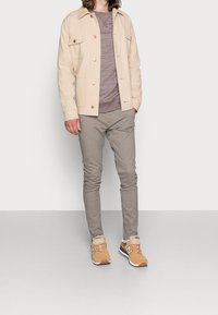 Giacca beige con bottone, camicia grigia a maniche lunghe, pantaloni grigi chiari e sneakers beige con accenti marroni. I dettagli includono tasche laterali e maniche a risvolto.