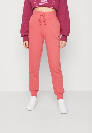 Persoon die koraalroze Nike joggingbroek met trekkoord draagt, gecombineerd met een cropped bordeauxrode Nike trui en witte sneakers.
