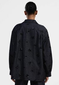 Camicia in denim scuro con una vestibilità ampia, caratterizzata da motivi floreali neri in paillettes distribuiti in modo casuale e un colletto classico. Maniche lunghe con polsini abbottonati.