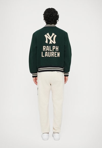 Bărbat cu spatele, îmbrăcat într-o jachetă varsity Ralph Lauren de culoare verde închis, cu logo alb, și pantaloni de trening crem, pe un fundal alb simplu.