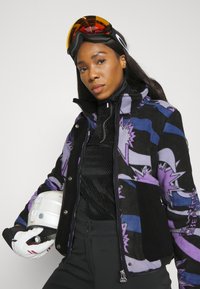 Multikleuren skijas met gedurfde paars, zwart en blauwe patronen, voorzien van zilverkleurige accenten, een hoge kraag en een gestructureerde zwarte basislaag.