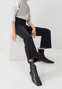 Black flared jeans with frayed hem paired with pointed-toe black leather ankle boots; model wearing a light textured top, seated on a white cube.
