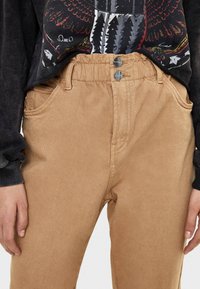 Pantalones de pana beige con cintura alta, que cuentan con una cinturilla fruncida, dos bolsillos frontales y cierre de doble botón. Textura suave.