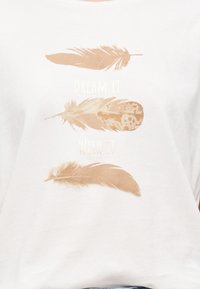 Τρία μπεζ φτερά με τις λέξεις "Ονειρέψου το" και "Εύχομαι το" τυπωμένες σε λευκή υφασμάτινη επιφάνεια.