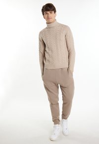 Beige kábelmintás garbós pulóver bézs joggerrel párosítva, oldalsó zsebekkel, fehér sneakerrel, egyszínű háttér előtt.
