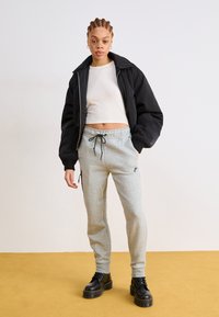 Veste bomber noire, crop top blanc ajusté, pantalon de jogging gris avec logo, et bottes plateformes noires. Tenue décontractée avec des textures contrastées.