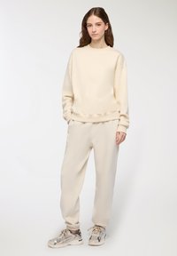 Felpa di colore crema, morbida, con vestibilità rilassata e polsini a costine, abbinata a pantaloni della tuta tapered coordinati. Indossata con sneakers beige.