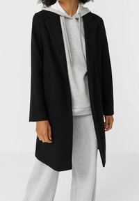 Manteau en laine noire à coupe droite, manches longues et deux poches latérales, porté sur un sweat à capuche gris clair et un pantalon de jogging gris assorti.