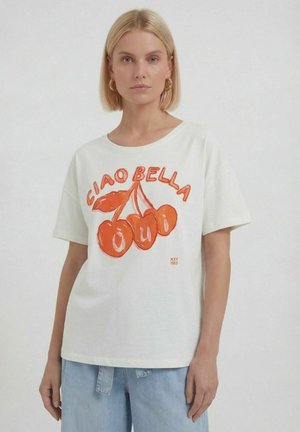 Blondine vrouw draagt een wit T-shirt met oranje kersen en de tekst "Ciao Bella" en lichtblauwe spijkerbroek tegen een effen achtergrond.