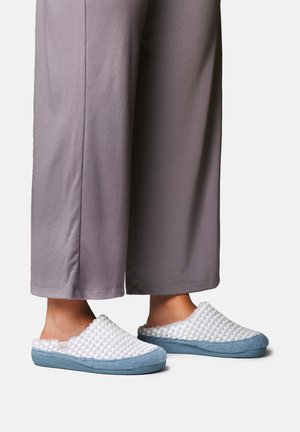 Fødder iført lyseblå og hvide teksturerede slip-on tøfler med såler, kombineret med grå bukser med vide ben.