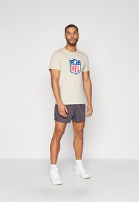 Bež majica s NFL logom s plavim i crvenim detaljima, uparena s lako sivim kratkim hlačama i bijelim tenisicama. Model stoji u neutralnom okruženju.
