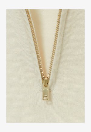 Grietai spalvos megzta medžiaga turi centrinius auksinius užtrauktukus, pabrėždama jos glotnią tekstūrą ir tiksliai siuvinėtas briaunas.